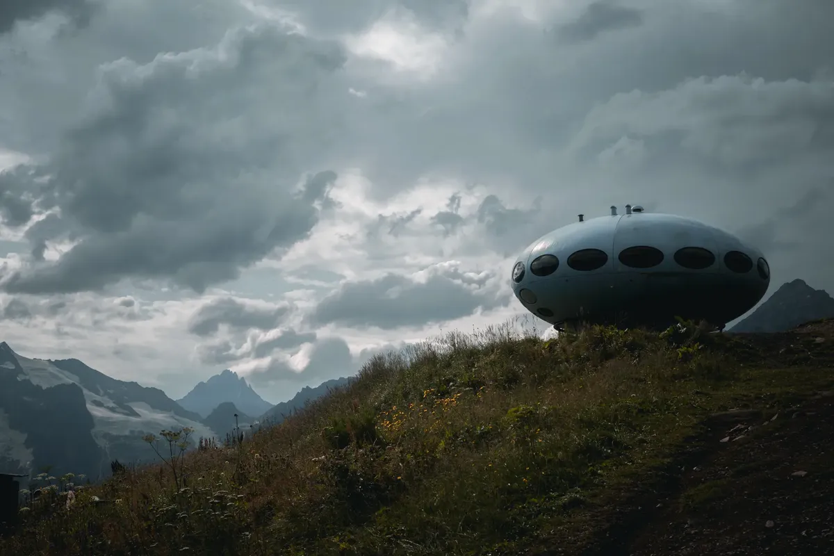 Unique UFO-like structure on a grassy hill with a cloudy mountain backdrop.