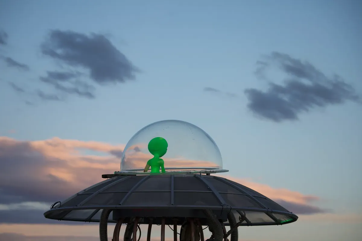 A playful alien figure inside a UFO replica against a vibrant evening sky.